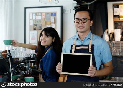 Young asian barista holding blank chalkboard with smiling face at cafe counter background, food and drink concept