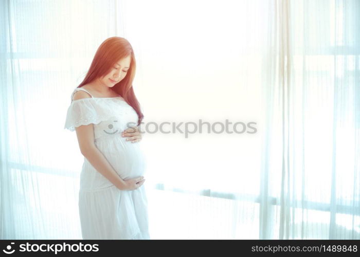 Young Asian beautiful pregnant woman standing near window at home