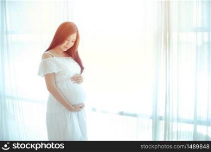 Young Asian beautiful pregnant woman standing near window at home