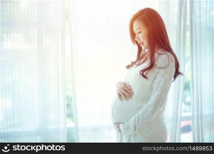 Young Asian beautiful pregnant woman standing near window at home