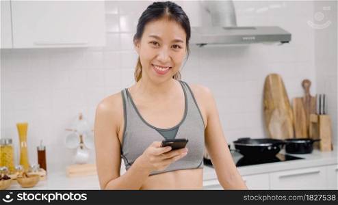 Young Asian blogger woman using smartphone for talking, chatting and checking social media in the kitchen, female in sport clothing cooking at home. Lifestyle women relax at home concept.
