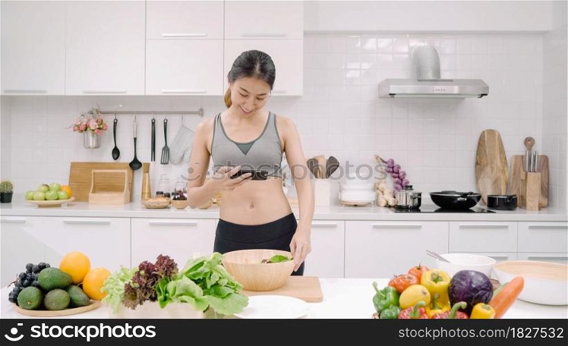 Young Asian blogger woman using smartphone photo post in social media in the kitchen, female in sport clothing making salad at home. Lifestyle women relax at home concept.
