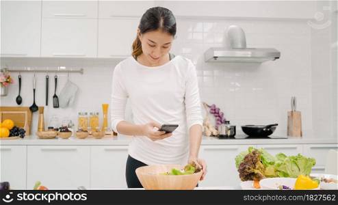 Young Asian blogger woman using smartphone photo post in social media in the kitchen, female making salad at home. Lifestyle women relax at home concept.