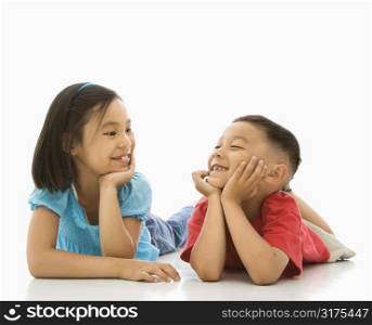Young Asian brother and sister lying on floor with head on hands looking at eachother smiling.