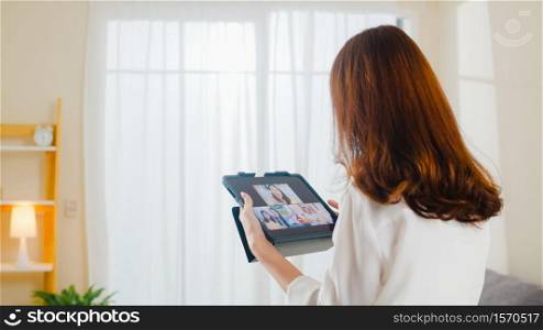 Young Asian business female using tablet video call talking with family while working from home at living room. Self-isolation, social distancing, quarantine for coronavirus in next normal concept.
