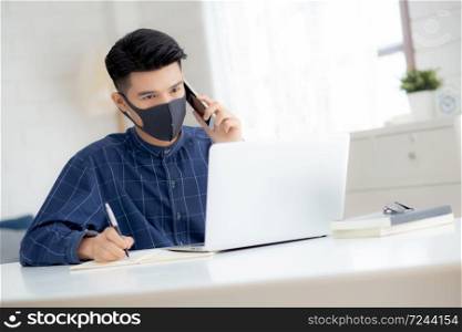 Young asian business man in face mask talking smartphone and work on laptop computer for protect covid-19, businessman quarantine work from home and writing notebook, social distancing and stay home.