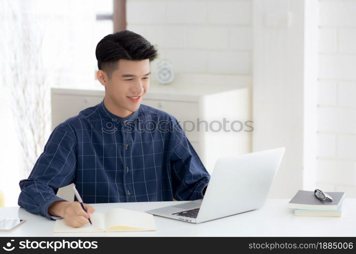 Young asian business man writing on notebook for planning working and using laptop computer on desk at home, notes about finance, male study and learning, business and communication concept.