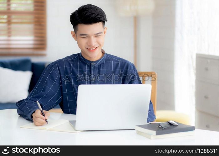 Young asian business man writing on notebook for planning working and using laptop computer on desk at home, notes about finance, male study and learning, business and communication concept.
