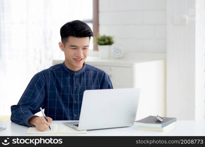 Young asian business man writing on notebook for planning working and using laptop computer on desk at home, notes about finance, male study and learning, business and communication concept.