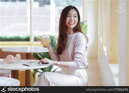 Young Asian business woman in casual clothes smile and sitting in cafe holding a glass of juice and looking out the window with tablet and pink headphones on the table