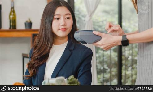 Young Asian business women pay contactless at coffee shop. Asian happy girl barista waiter wear gray apron holding credit card reader machine for customer using mobile phone scan pay in cafe.
