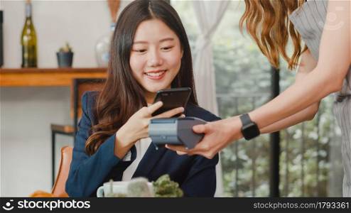 Young Asian business women pay contactless at coffee shop. Asian happy girl barista waiter wear gray apron holding credit card reader machine for customer using mobile phone scan pay in cafe.