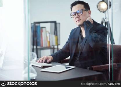 Young Asian businessman feeling tired while sitting at his working place and massaging his neck .