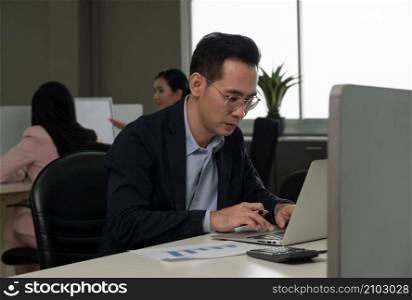 Young Asian businessman in eyeglasses working on laptop at his desk in open space office with colleagues in the background