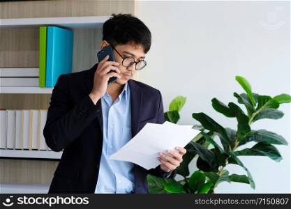 Young asian businessman reading papers talking phone at office, business communication and technology concept