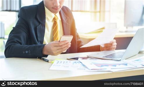 Young asian businessman sitting and working on desk and using smart phone connecting internet and documents at office, employee or company ceo using online apps software at workplace