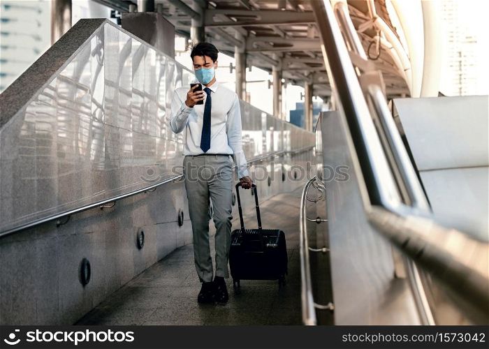 Young Asian Businessman Wearing a Surgical Mask and Using a Smart Phone while Walking with Suitcase in the Airport or Public Transportation Station.Healthcare in New Normal Lifestyle Concept