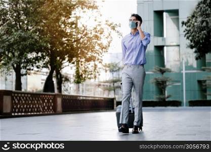Young Asian Businessman Wearing a Surgical Mask and Using a Smart Phone while Walking with Suitcase in the City.Healthcare in New Normal Lifestyle Concept