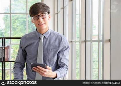 Young asian businessman wearing glasses smiling and looking at camera and holding a digital tablet PC in his hand while standing at the big window in office