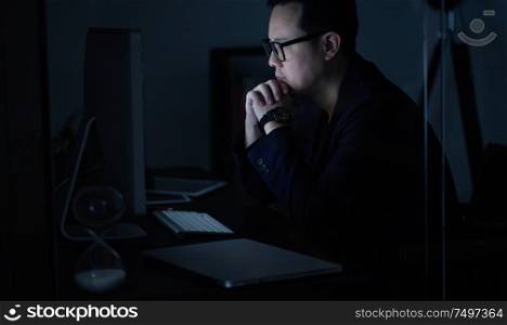Young Asian businessman working on a laptop while watching computer at his office desk at night .