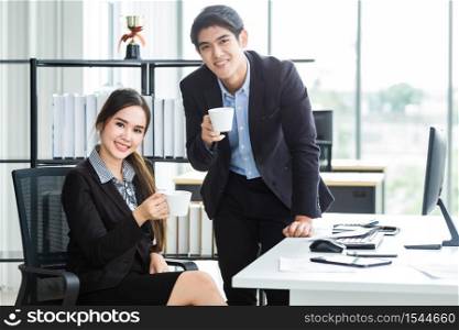 Young Asian businesswoman and businessman partners while working together and relax holding a cup of coffee in before work business meeting with computer in office background,Working couple