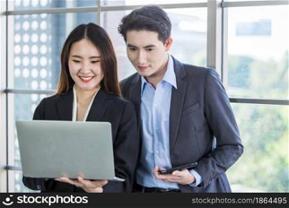 Young Asian businesswoman and businessman partners while working together with laptop computer and business man holding a tablet in office background,Working couple