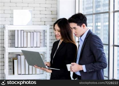 Young Asian businesswoman and businessman partners while working together with laptop computer and business man holding a cup of coffee in office background,Working couple