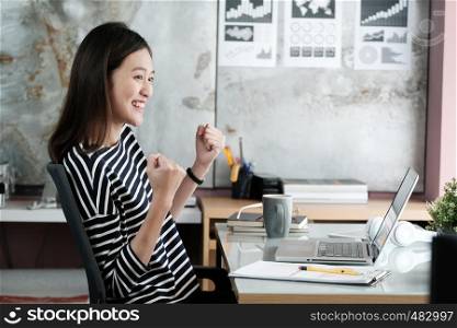 Young asian businesswoman arm up and smiling while sitting at her desk office, positive expression, sucess in business concept