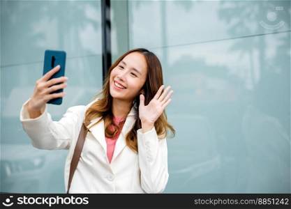 Young Asian businesswoman during video chat in smart phone outside in the city next the building. Happy woman enjoying sunlight outdoors. Lifestyle and technology.