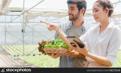 Young Asian businesswoman farmer hold tablet show performance of her organic farm to hispanic man buyer at greenhouse farm garden, New innovation technology in the cultivation concept.