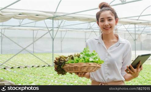 Young Asian businesswoman farmer with product selling good quality plant and vegetable and holding tablet looking at camera at organic farm in greenhouse garden, Farm innovation technology concept.