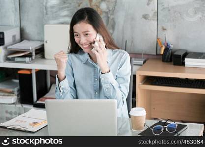 Young asian businesswoman talking phone and arms up with smiling face while working at her office desk, success in business concept