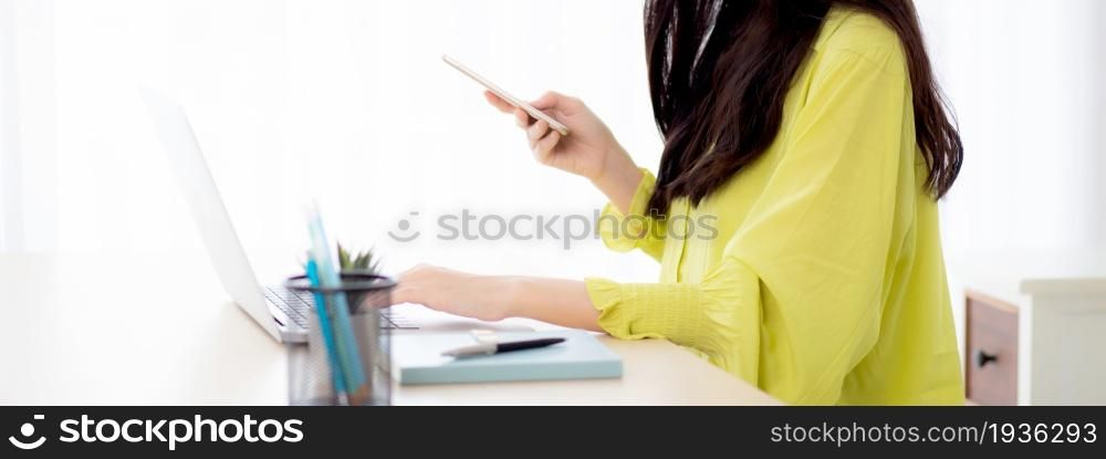 Young asian businesswoman working on laptop computer online on desk at home office, freelance woman looking message on smart phone and typing on notebook on table, business and communication concept.
