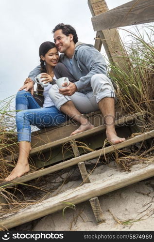 Young Asian Chinese man &amp; woman, boy &amp; girl, couple sitting on wooden steps overlooking a beach drinking mugs of tea or coffee