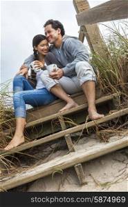 Young Asian Chinese man &amp; woman, boy &amp; girl, couple sitting on wooden steps overlooking a beach drinking mugs of tea or coffee