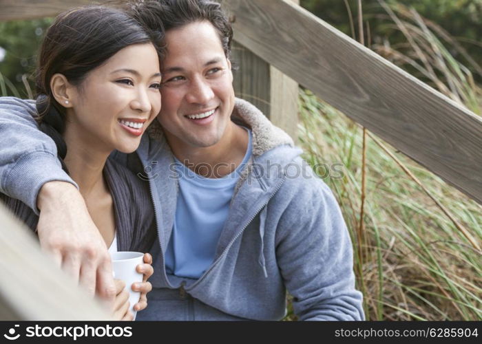 Young Asian Chinese man & woman, boy & girl, couple sitting on wooden steps overlooking a beach drinking mugs of tea or coffee