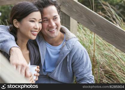 Young Asian Chinese man & woman, boy & girl, couple sitting on wooden steps overlooking a beach drinking mugs of tea or coffee