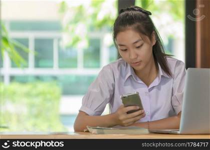 Young Asian college students in uniform beautiful girl relax and smile using smartphone and laptop computer at coffee shop for learning online, Casual business, social meeting, or education concept.. Education concept.