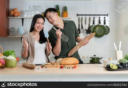 Young Asian couple having fun in kitchen, singing and dancing while cooking at home. Playful lover using spatulas and egg whisk as microphones and loaf stick of bread as guitar doing band together