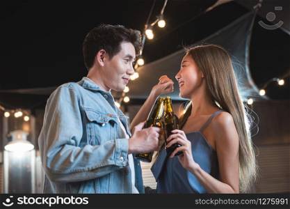 Young asian couple lover have fun dancing and drinking in night party on rooftop floor nightclub hand holding beer bottle and eye contact flirting at couple party.