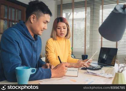 Young asian couple managing finances, reviewing their bank accounts using laptop computer and calculator at modern home. Woman and man doing paperwork together, paying taxes online on notebook pc.