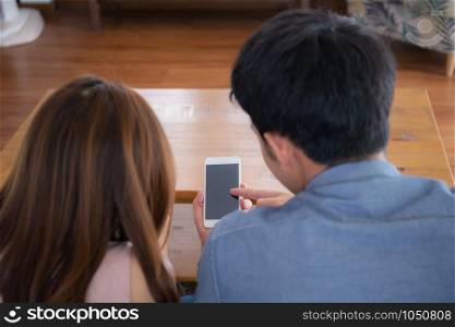 Young asian couple or friend looking smart mobile phone with enjoy at living room, happy family relax and fun media entertainment on smartphone blank together at home, lifestyle concept.