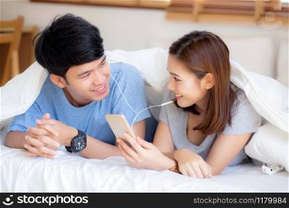 Young asian couple smile listen music with smart mobile phone on bed at bedroom with fun and enjoy, family sharing podcast or watching video from internet online with relax, lifestyle concept.