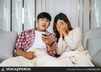 Young Asian couple spend time watch scary movie on TV in living room in afternoon, Young man and woman sit on sofa and cover with blanket while watching television together, copy space