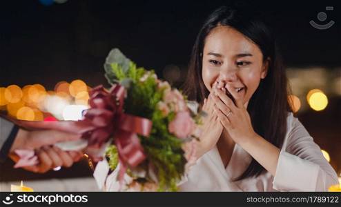 Young asian couple surprise moment with rose bouquet at romantic dinner in rooftop restaurant at city night with sweet moment celebrate anniversary. Love relationship, marry concept.