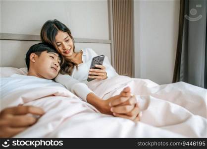 Young Asian couple using mobile phone while lying on bed and smiling with happiness together. Happy couple lifestyle concept