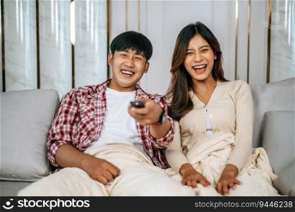 Young  Asian couple watch television on sofa in new house and watch funny movie together. They use remote to change  channel, Happy family Concept