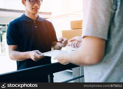 Young asian delivery staff holding the pen and documents submitting giving to the customer receiving the parcel at front house.