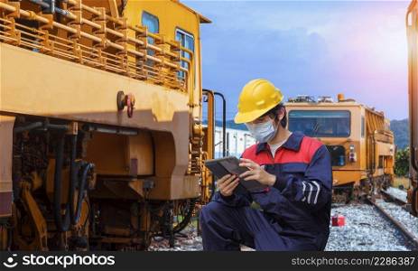 Young Asian engineer in safety workwear with protective mask using digital tablet to record information of the old trains during maintenance at locomotive maintenance station in countryside