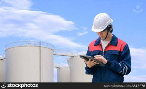 Young Asian engineer using digital tablet to working his job with blurred background of storage fuel tanks against cloud on blue sky in refinery base area